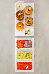 Four bagels with spreads are elegantly arranged on the Inspired Generations Acrylic Bagels On Board Tray in White, accompanied by three trays of sliced tomatoes, red onions, cucumbers, and smoked salmon—each with convenient serving utensils—all set atop a light marble surface to create the perfect brunch board.