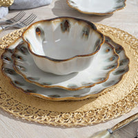 The Amoria Dinnerware set by Blue Pheasant—featuring a scalloped ceramic bowl and plate with brown and green accents—rests on a woven placemat atop a light wooden table, with cream-handled fork and knife beside it.