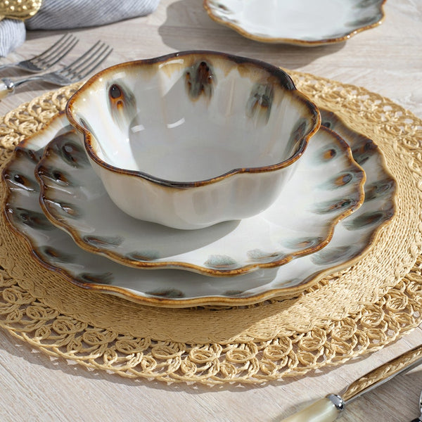 The Amoria Dinnerware set by Blue Pheasant—featuring a scalloped ceramic bowl and plate with brown and green accents—rests on a woven placemat atop a light wooden table, with cream-handled fork and knife beside it.