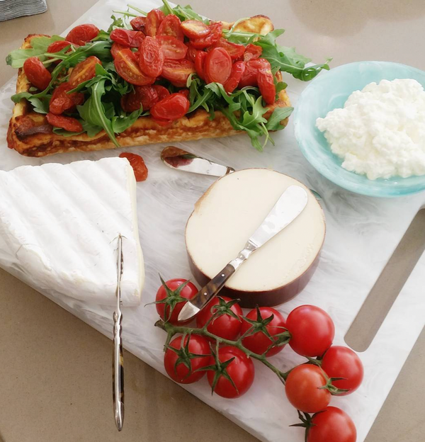 The Nashi Home Swirl Resin Rectangle Chopping Board White – Large displays a wedge of brie, smoked cheese, cottage cheese, vine tomatoes, and toast with arugula and roasted tomatoes. Cheese knives are set beside the cheeses for serving.