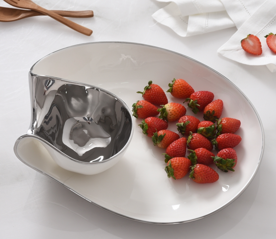 The PAMPA BAY Thin and Simple Chip & Dip is a modern white and silver porcelain serving platter with a separate silver compartment, holding fresh strawberries. Oven and dishwasher safe, it sits on a white surface with a napkin and sliced strawberries nearby.