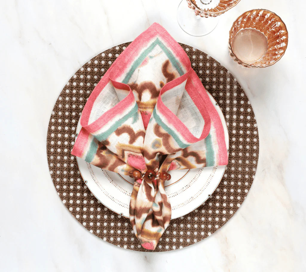 A decorative table setting features a patterned napkin in the Kim Seybert Bois Napkin Ring Set of 4 - Brown & Gold on a white plate, atop a brown woven placemat with two amber glasses on a white marble surface.