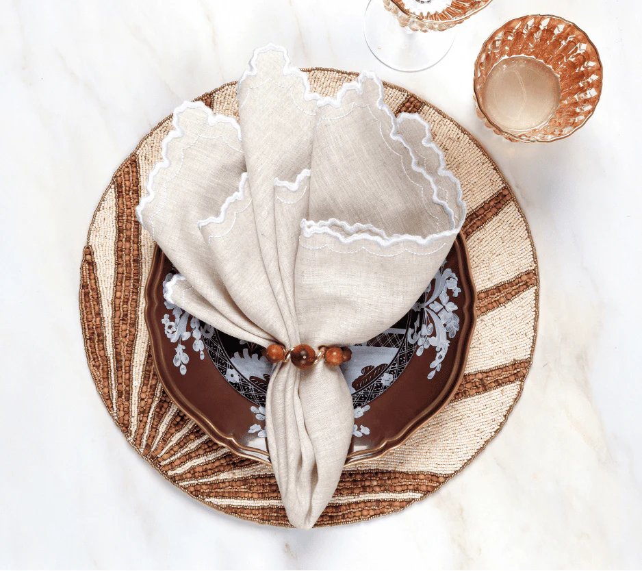 A table setting with a brown and white patterned plate, woven placemat, beige scalloped linen napkin held by the Kim Seybert Bois Napkin Ring Set of 4 - Brown & Gold, on marble beside two amber glasses.