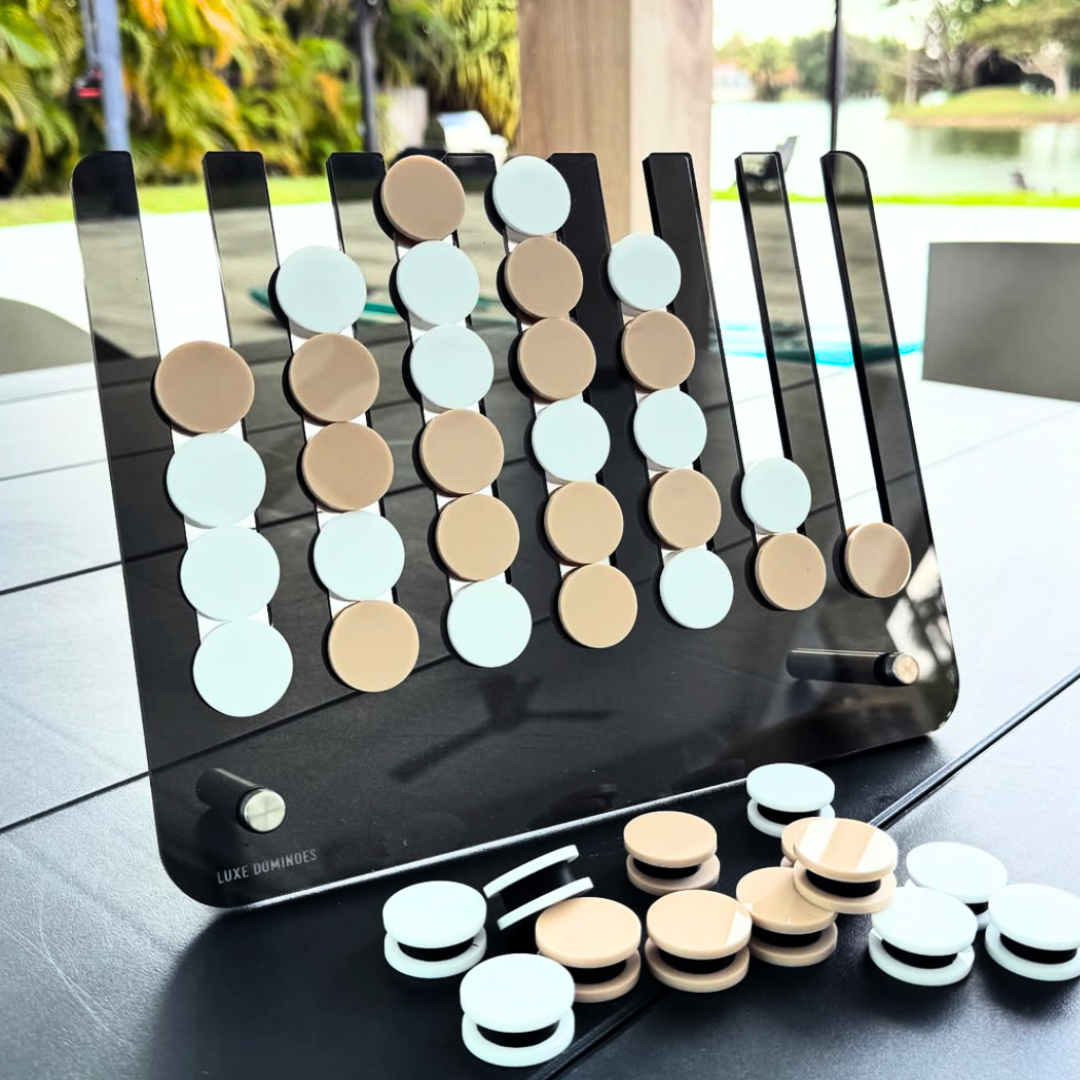 The Connect Four El Cuatro game by Luxe Dominoes, featuring chic beige and white pieces, is elegantly arranged on a black table outdoors. Extra pieces are on hand, while the backdrop of trees, water, and patio furniture creates an inviting atmosphere—perfect for challenging friends to see who can get four in a row first.