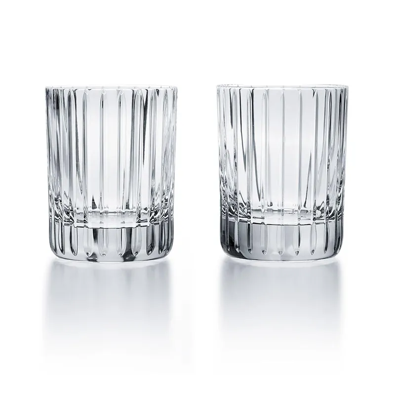 Two clear Baccarat Harmonie highball glasses with vertical ridges stand side by side on a reflective white surface, showcasing the modern design of this Baccarat barware set of 2.