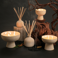 The Spiritual Diffuser by Blueme and beige ceramic candles sit on black pebbles and rustic wood against a dark backdrop. Lit candles release a calming aroma, creating a soothing, spa-like ambiance.