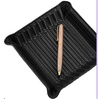 A gold and brown pen rests diagonally inside the Graphic Image Leather Valet Tray - Black Woven, featuring raised corners and a square design, set against a white background.