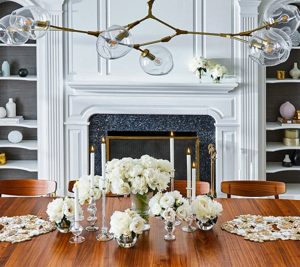 An elegant dining room features a wooden table adorned with white flowers and tall candles placed in Kim Seybert’s Ripple Candle Holder - Clear, whose iridescent luster adds a touch of refined beauty. A modern chandelier hangs overhead, while a white fireplace with decorative shelves forms the backdrop.