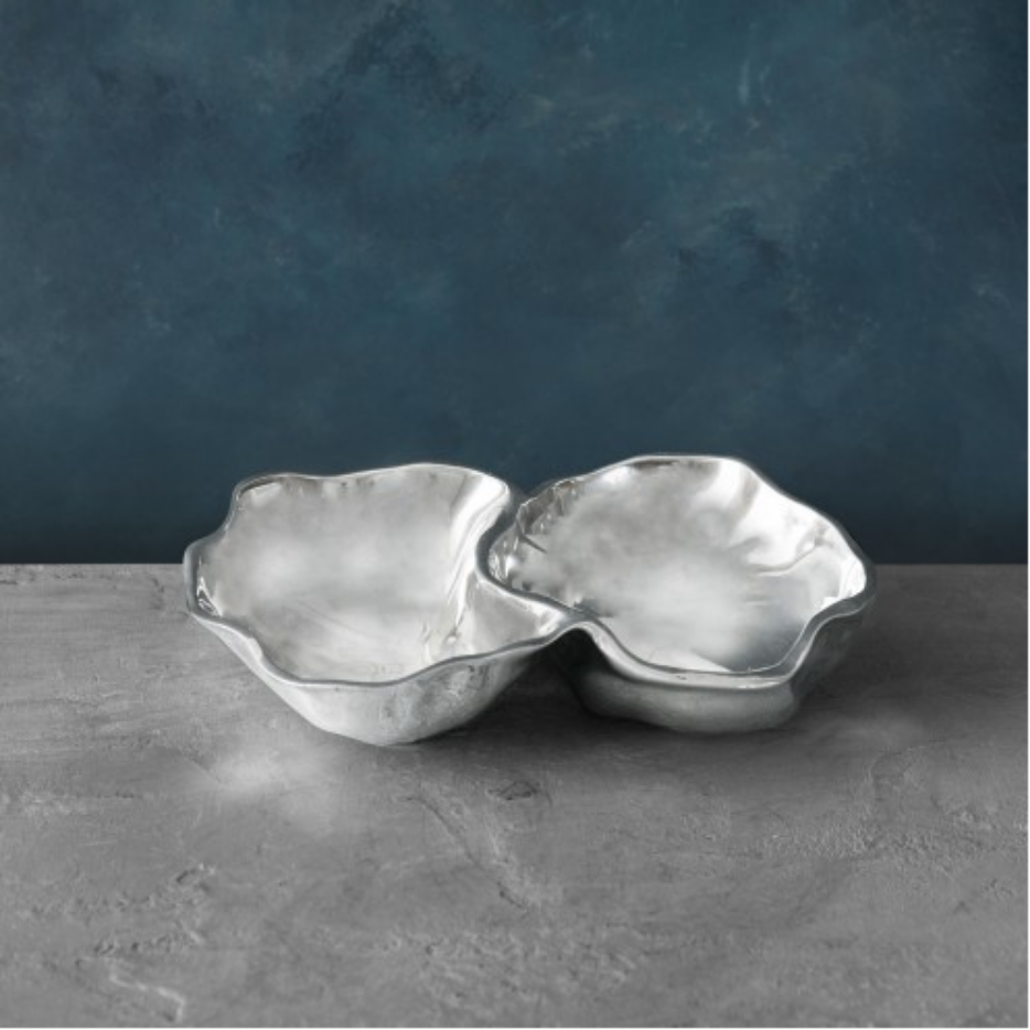 The Beatriz Ball Soho Onyx Double Dip Serving Bowl features a silver-toned, wavy-edged design—ideal for serving dips. Displayed on a gray textured surface with a blue textured background, it combines elegance and function.