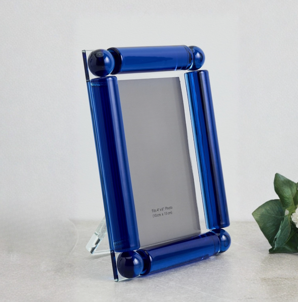 The Tizo Corner Ball Crystal Frame Blue features thick, cylindrical blue glass borders with crystal ball accents. It sits on a light surface with a green plant to the right, and its modern design is empty except for a size label.