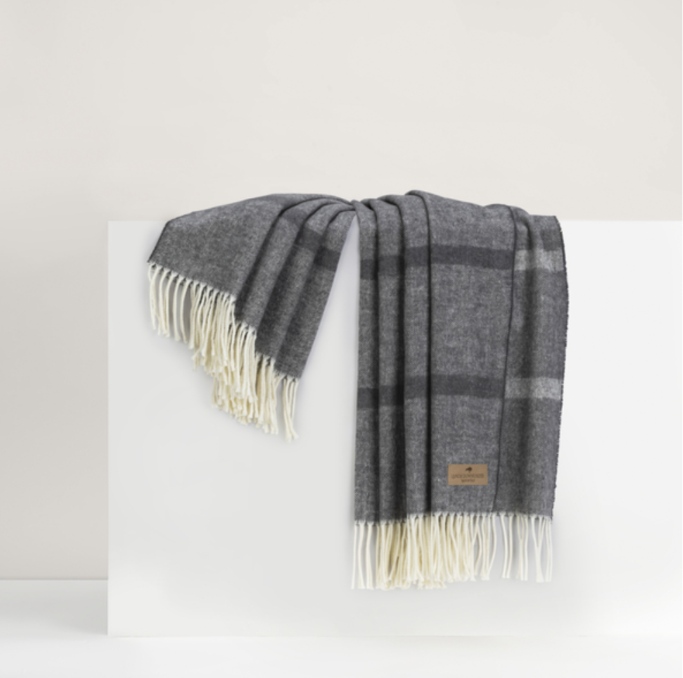 A charcoal gray Montauk Stripe Throw by Lands Downunder, featuring a subtle plaid pattern, cream fringed edges, and a small brown leather tag, is draped over a white surface. Expertly crafted in Italy.