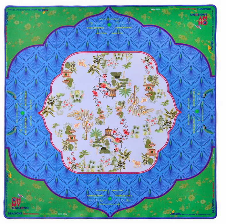 The Oh My Mahjong "Mahjong Mats" silk scarf features a central white area with small Asian-style houses, trees, and flowers, bordered by blue scalloped patterns and a green edge with decorative details and text.