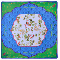 The Oh My Mahjong "Mahjong Mats" silk scarf features a central white area with small Asian-style houses, trees, and flowers, bordered by blue scalloped patterns and a green edge with decorative details and text.