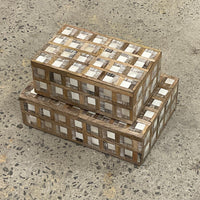 Two Plaid Wood and Bone Boxes from Bojay, featuring a bold checkerboard pattern crafted from various wood shades, are stacked on a speckled concrete floor. These decorative boxes combine masculine styling with rustic appeal, providing both functional storage and distinctive design to any space.