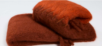 Two fluffy, rust-colored Stitching Throw blankets by Mantas Ezcaray are shown—one flat, one rolled with decorative leather ties. Both have a soft, fuzzy texture and capture the luxury, handmade tradition of Ezcaray.