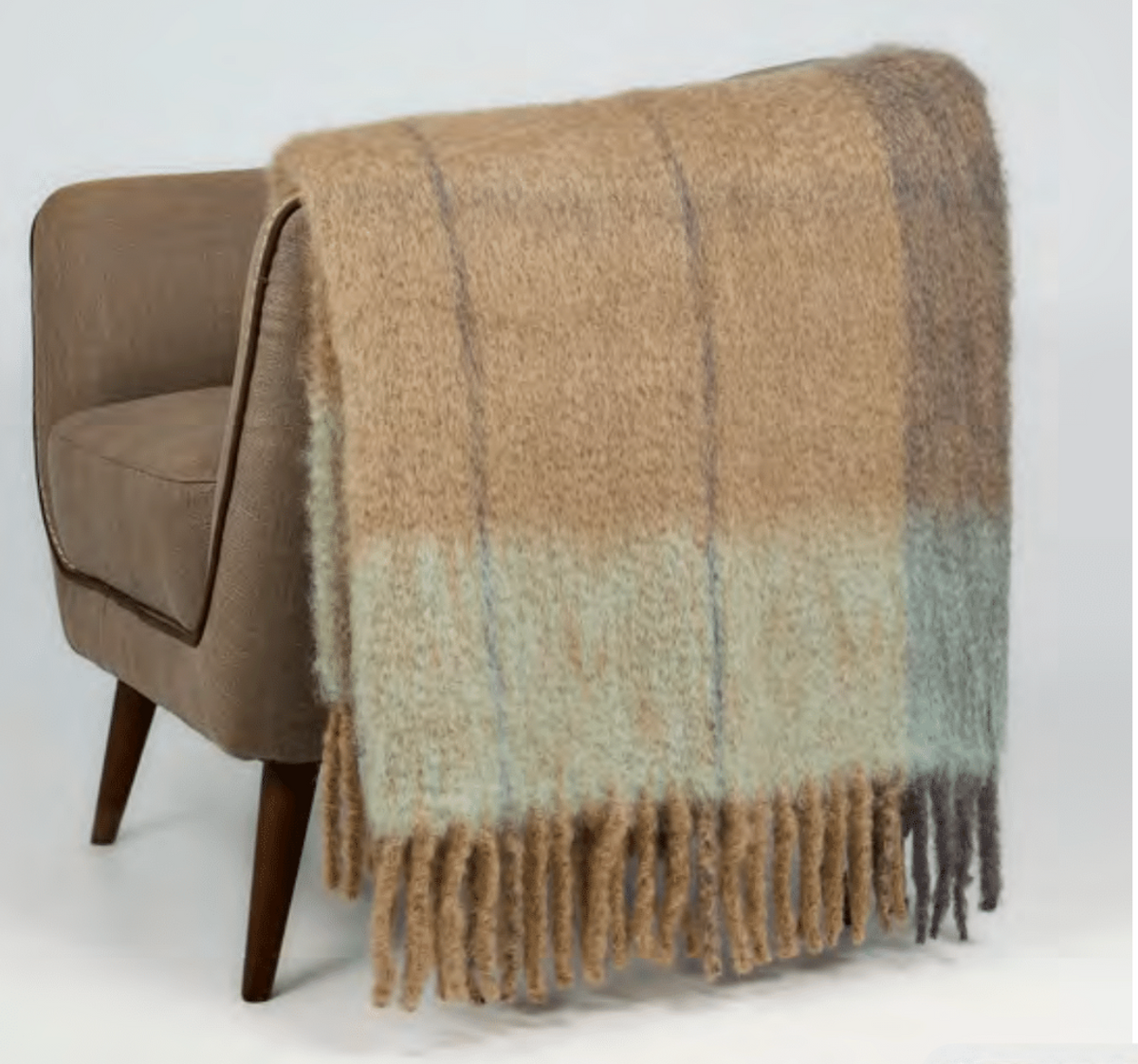 The Mantas Ezcaray Twist Frame Throw, featuring beige, light blue, and light brown stripes with long fringe, is draped over a mid-century modern tan chair. Expertly handcrafted in Spain for exceptional quality.