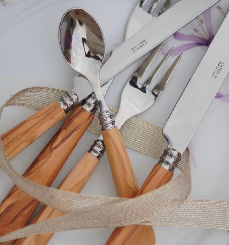 The Capdeco Altea Flatware 5 Piece Setting in Olive Wood, featuring knives, forks, and a spoon, is displayed on a white plate with a beige ribbon elegantly draped around the pieces.