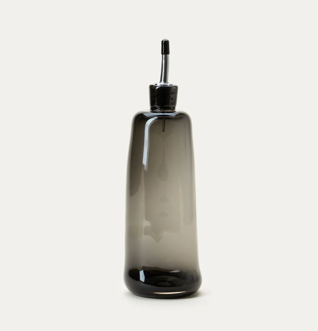 The Handblown Oil Cruet by Gary Bodker features a smoky gray glass design with a tapered silhouette and a metal pour spout, highlighting exceptional craftsmanship. Photographed against a plain white background, this elegant kitchen accessory brings both style and function to your space.