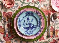 A vibrant table setting features a green charger, pink scalloped plate, and the Ginori 1735 Oriente Italiano Dinnerware Iris plate with chinoiserie florals, all set with silver utensils, glasses, and a floral tablecloth.