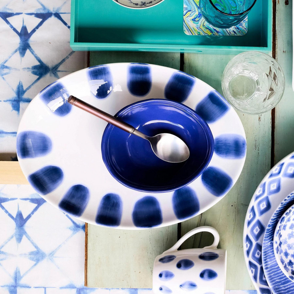 The Vietri Santorini Dot Small Oval Platter holds a blue bowl with a spoon. Surrounding it are a patterned mug, glass cup, and other blue and white dishes arranged on a rustic wooden surface.