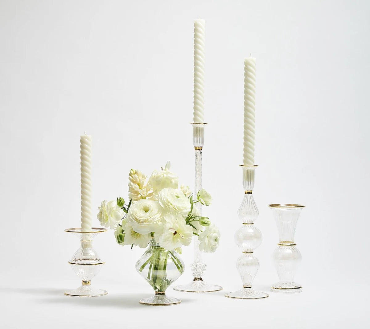 A group of clear glass candlesticks with white candles and a matching Kim Seybert Scallop Bud Vase in clear, elegantly holding white flowers—all featuring gold trim—are displayed on a white background.