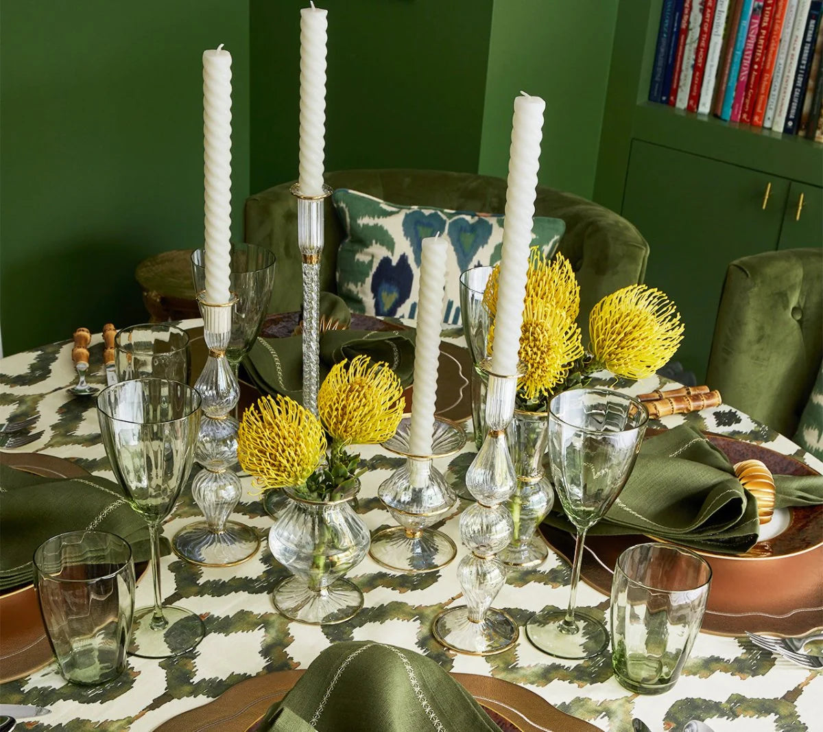 Elegant table set with green napkins, a patterned tablecloth, clear mouth-blown glassware, white candles in glass holders, and yellow flowers arranged in hand-crafted vases—including the Kim Seybert Scallop Bud Vase - Clear. Books are visible on a green shelf in the background.