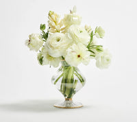 A bouquet of white flowers, including ranunculus and other blooms with green stems visible in the water, is arranged in the hand-crafted Scallop Bud Vase - Clear by Kim Seybert, set against a plain white background.
