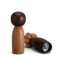 The Picanto Salt + Pepper Mills Set by L'Objet features two mills—one upright with a dark top and light oak body, the other showing its metal grinder and dark wood finish.