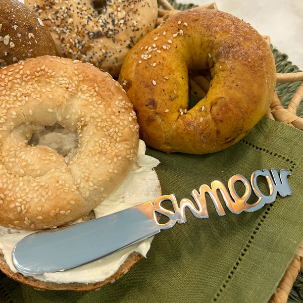 A sesame bagel with cream cheese, surrounded by other bagels on a tray, sits atop a green napkin. Next to the bagels lies an Inspired Generations Schmear Spreader knife, featuring the word “smear” cut into its handle.