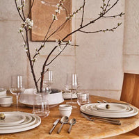 The table features a Serax Dune Oval Platter Alabaster - 18", set with white plates, silver cutlery, and clear glassware. A vase with flowering branches completes the elegant, minimalist setting in neutral tones.