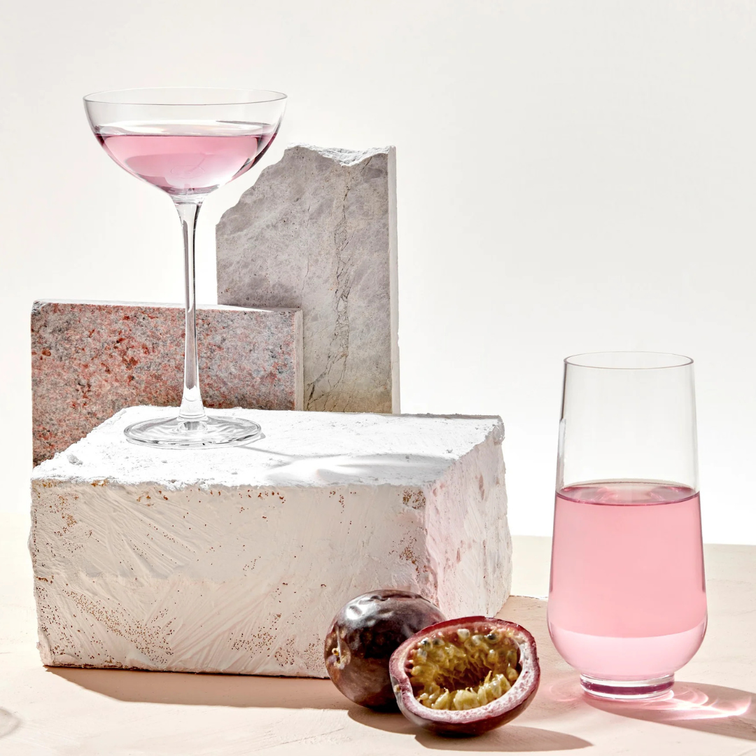 Two glasses from the Hepburn Glass Set of 4 by Nude Glassware, both filled with light pink liquid, are displayed on and beside white stone blocks. Whole and halved passion fruit sit in front against a minimalist, bright background.
