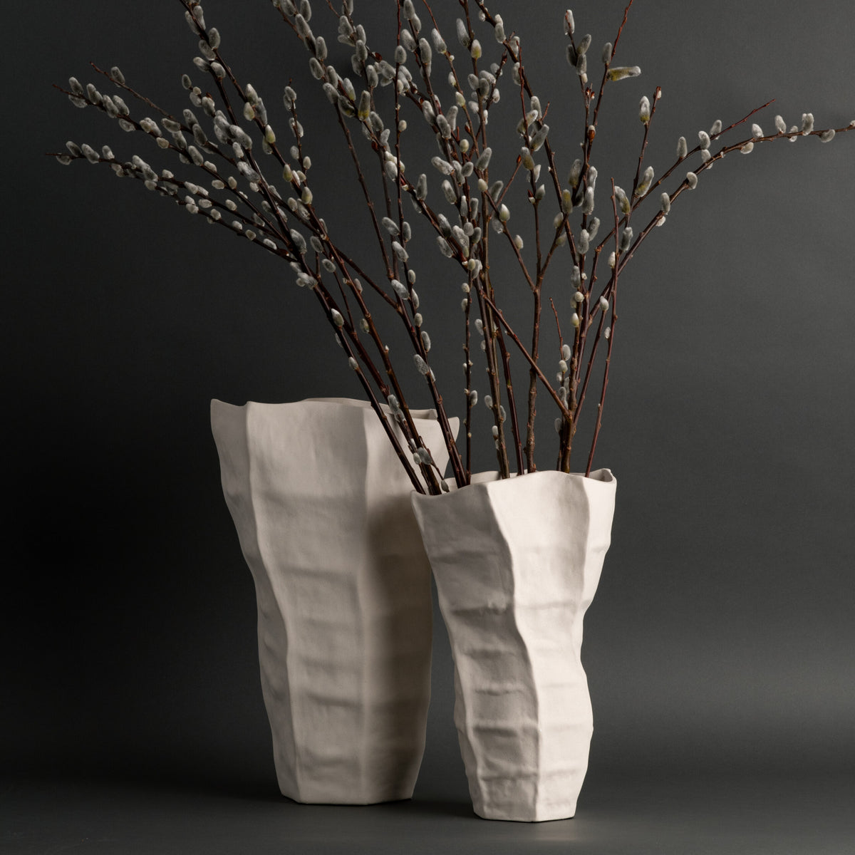 Two tall, off-white Lucille Vases by FlowDecor Inc., featuring textured, angular ceramic forms, hold long, slender branches of pussy willow against a dark gray background. Their artisanal quality and sculptural design from FlowDecor Inc. add an elegant touch to the composition.