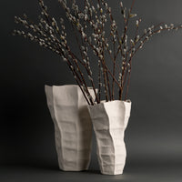 Two tall, off-white Lucille Vases by FlowDecor Inc., featuring textured, angular ceramic forms, hold long, slender branches of pussy willow against a dark gray background. Their artisanal quality and sculptural design from FlowDecor Inc. add an elegant touch to the composition.