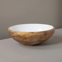 The Be Home Mayan Wood Footed Bowl (17"D) is handcrafted in India from sustainably sourced wood, featuring a natural grain exterior and glossy white interior. It rests on a light gray surface with a plain gray background.