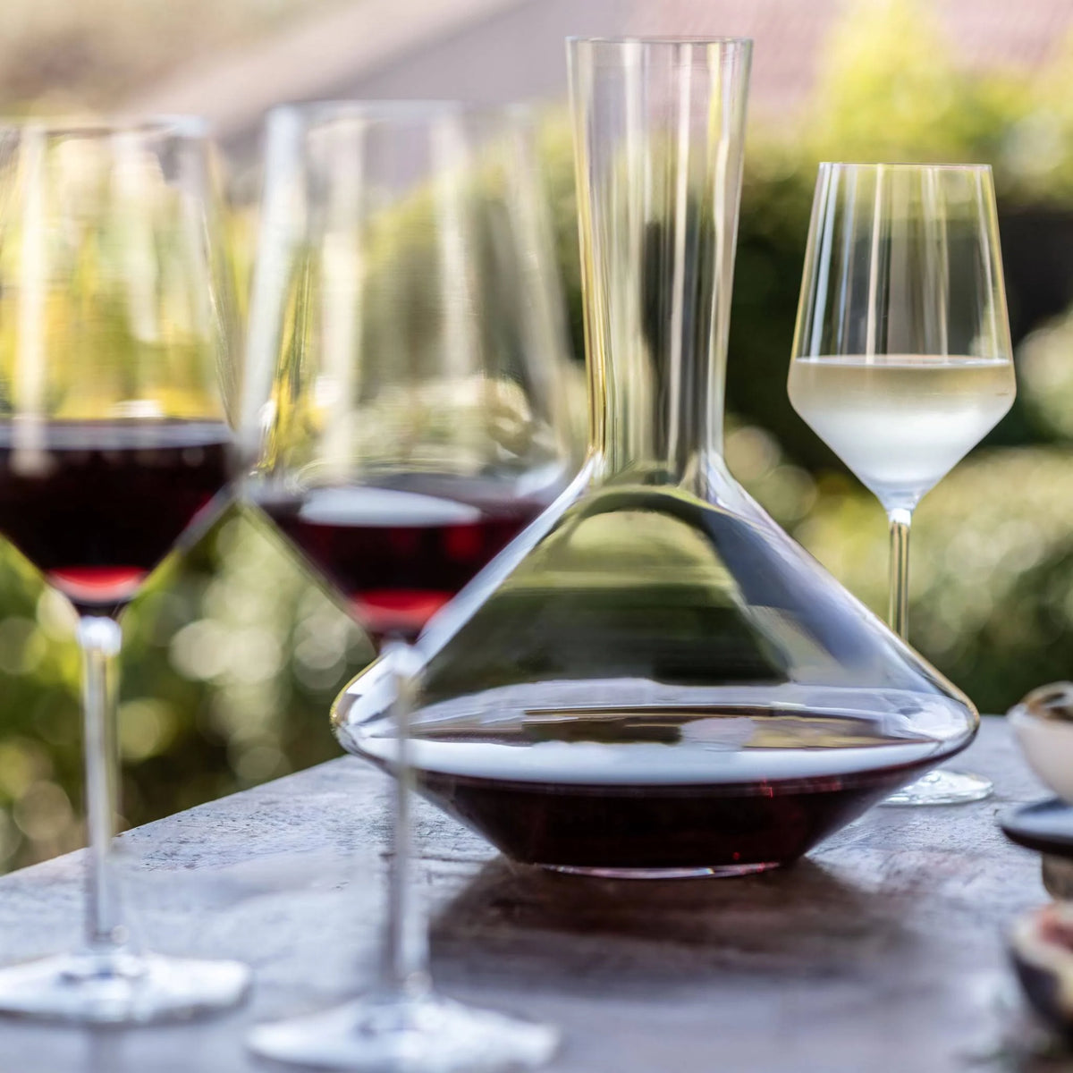 Three wine glasses—two with red, one with white—sit beside the Zwiesel Fortessa Zweisel Pure Red Wine Decanter on an outdoor table made of Tritan Crystal Glass, with blurred greenery in the background.