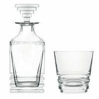The Saint-Louis Oxymore Drinkware set features a clear glass decanter with a geometric, stepped stopper beside a matching tumbler, both with angular stacked bases, exemplifying contemporary French glassware style.