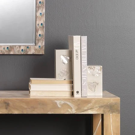 A wooden console table with stacked books, upright books held by two elegant Jamie Young Stone Bookends Pearl Set of 2, and part of a wall mirror with a decorative peacock feather frame, against a gray wall.