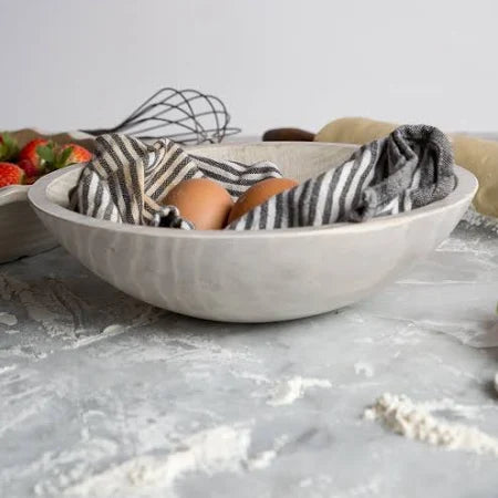 The Peterman Round Wood Bowl Driftwood 15" cradles two brown eggs in a black and white striped cloth, set on a floured counter with baking utensils and strawberries in the background.