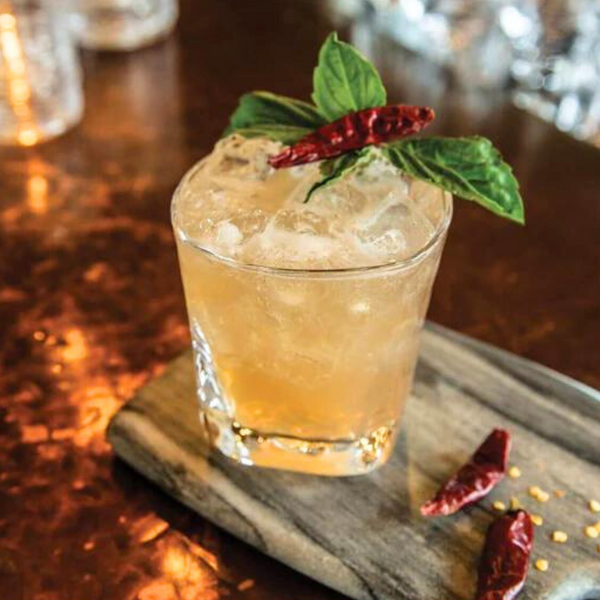 A cocktail with ice, garnished with basil and a dried red chili, is served in a Simon Pearce Woodbury DOF glass on a wooden board with extra chilies atop a copper-toned surface.