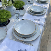 Elegant outdoor table setting featuring Molecot's Simply Gold Dinnerware, silver cutlery, embroidered napkins, water glasses, name cards, and green floral centerpieces on a white tablecloth.