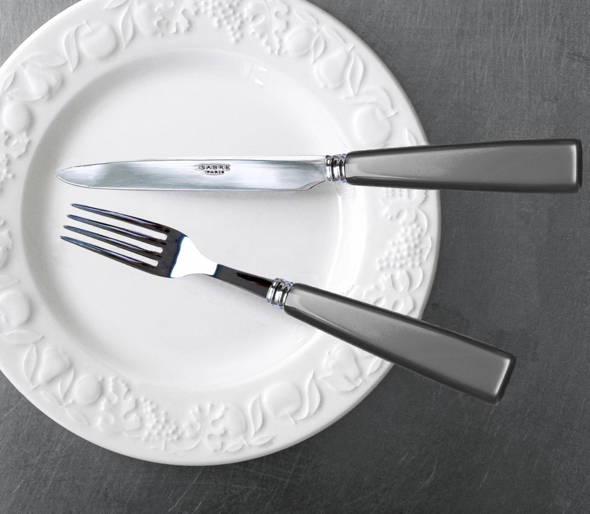 A white ceramic plate with embossed fruit designs holds Sabre’s Icone Flatware 5 Piece Setting—18/10 stainless steel fork and knife with dark handles—angled on a dark gray surface, ideal for any icon collection.