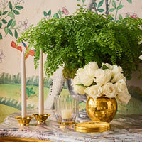 A marble table features two white candles in Aerin’s Olivia Taper Candleholders, a glass votive candle, a gold vase filled with white roses, and a green plant, set before floral and bird Art Deco-inspired wallpaper.