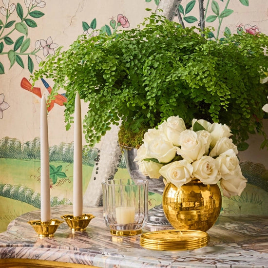 A marble table features two white candles in Aerin’s Olivia Taper Candleholders, a glass votive candle, a gold vase filled with white roses, and a green plant, set before floral and bird Art Deco-inspired wallpaper.