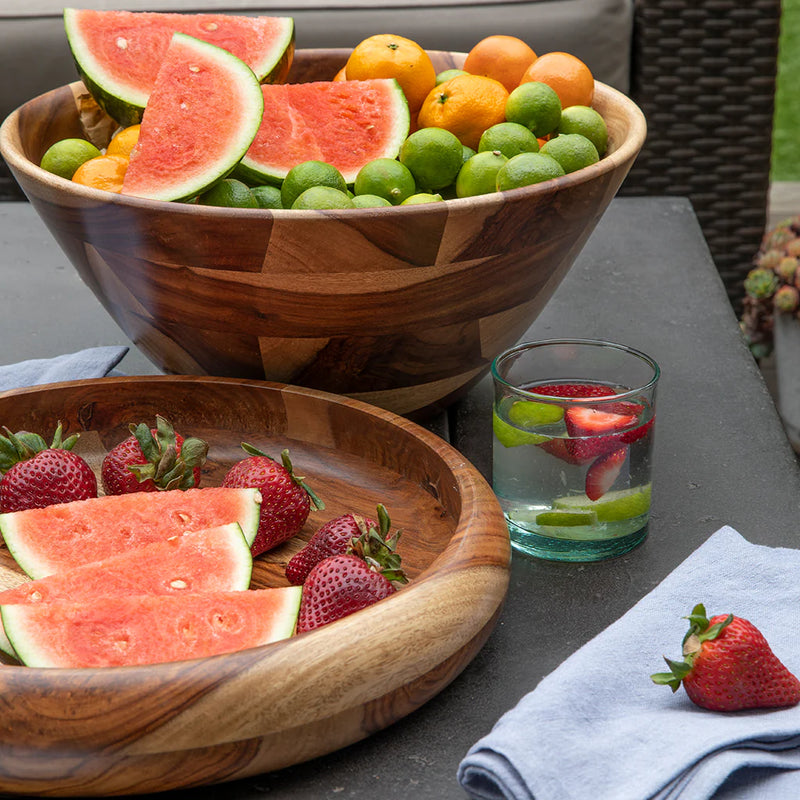 The Be Home Bumi Rosewood Serving Bowl holds watermelon, oranges, and limes beside a plate of strawberries and watermelon. A fruit-infused water glass and a blue napkin complete the setting on the dark outdoor table.