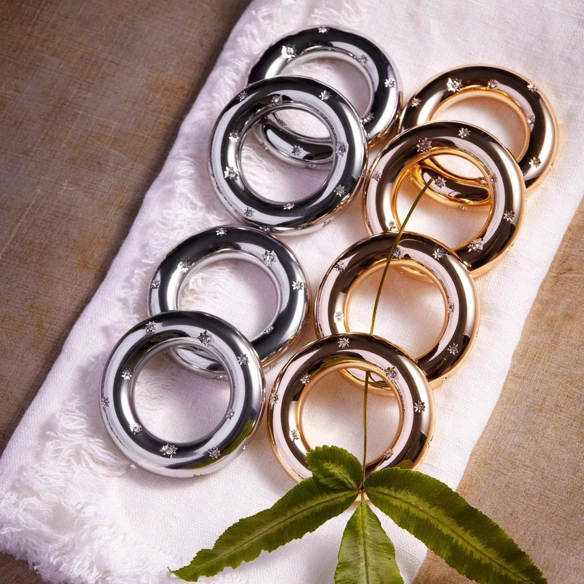 The L'Objet Stars Napkin Jewels Set of 4 features shiny silver and 24k gold plated rings with elegant details, beautifully arranged on a white cloth and topped with a green leafy stem.