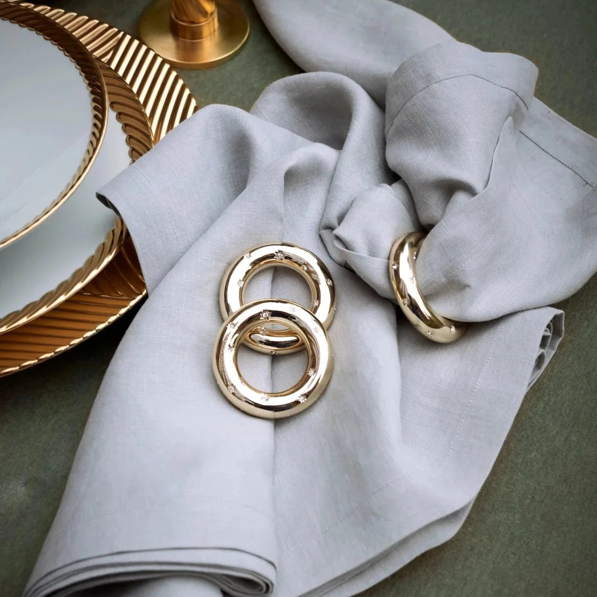 A close-up of a gray linen napkin with two L'Objet Stars Napkin Jewels (Set of 4, 24k gold plated), beside white plates with gold rims on a green tablecloth. A gold candlestick appears in the background.