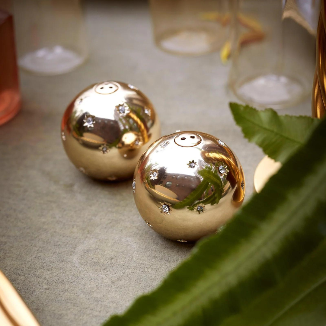Two gleaming Stars Spice Jewels Salt & Pepper Set - Platinum shakers by L'Objet, with small star accents, rest on a gray surface near green leaves and glassware.