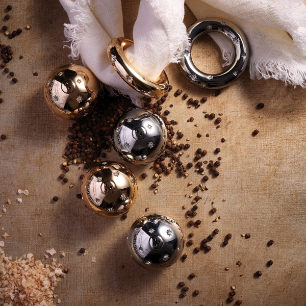 Platinum-plated spherical and ring-shaped objects from the Stars Spice Jewels Salt & Pepper Set by L'Objet are displayed on a textured surface with black peppercorns, coarse salt, and white fabric.