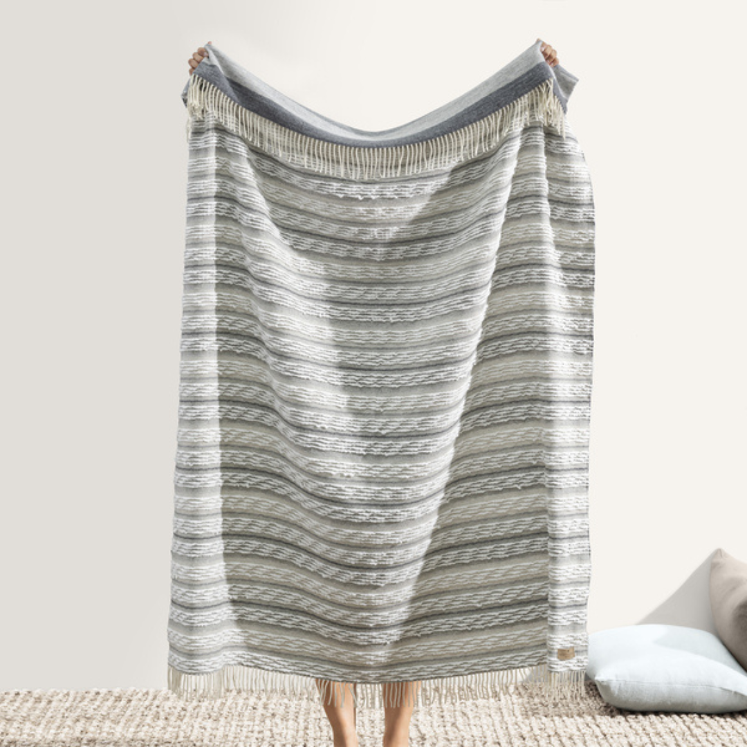 A person holds up the large Strato Throw by Lands Downunder, covering most of their body with only hands and feet visible. The simple setting includes a woven rug and two cushions on the floor.
