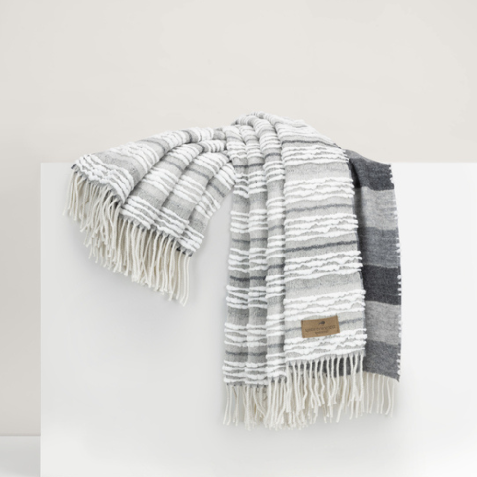 The Strato Throw by Lands Downunder, featuring gray and white stripes and fringe on both ends, is draped over a white surface. A small brown tag appears on one side of this reversible throw blanket.